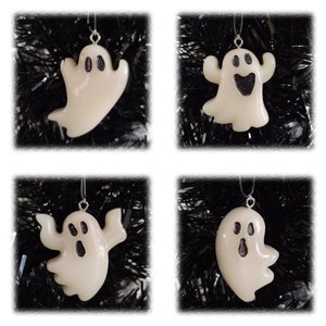 May include: Four white ghost ornaments with black glitter accents, hanging against a black background. Each ghost has a unique expression, perfect for Halloween decorations. The ornaments are suspended by silver strings.