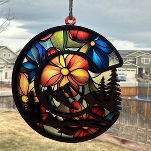 May include: A round wooden ornament with a stained glass design of colorful flowers and a mountain silhouette. The ornament is hanging from a string.