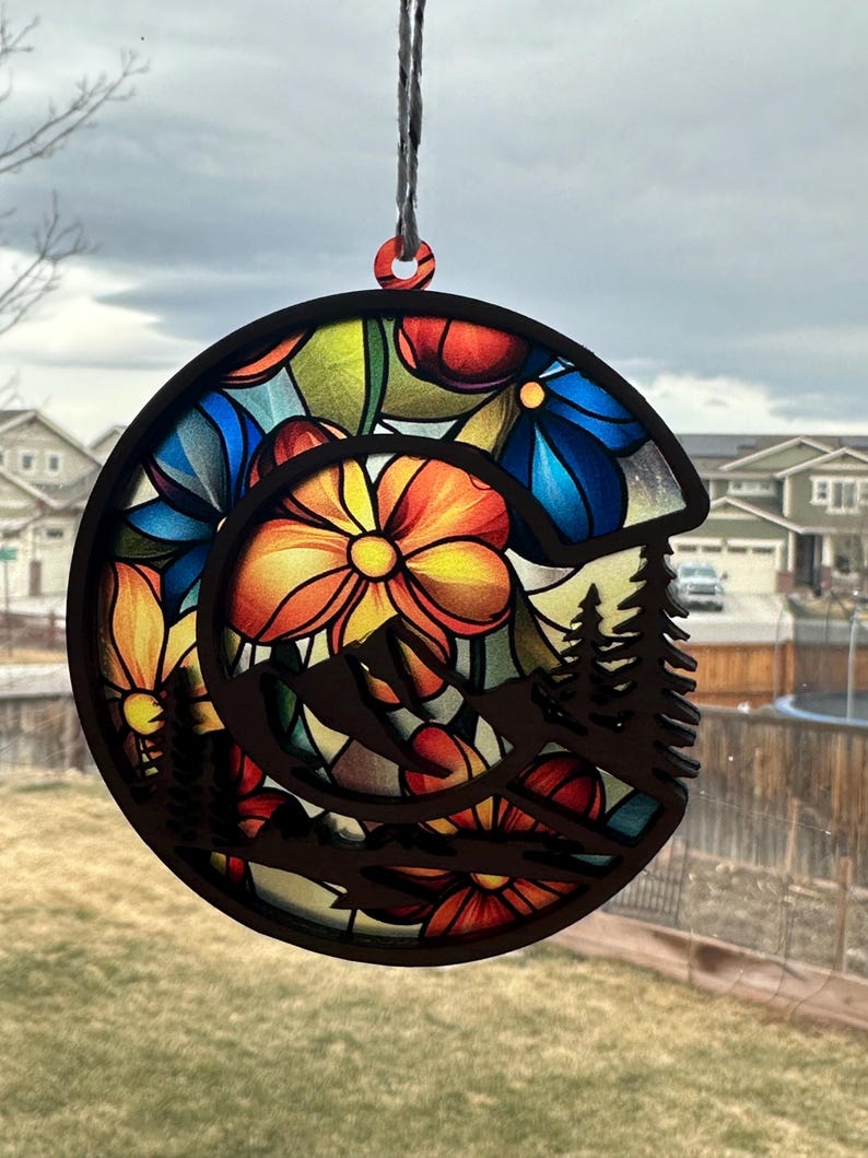 May include: A round wooden ornament with a stained glass design of colorful flowers and a mountain silhouette. The ornament is hanging from a string.