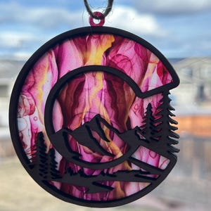 May include: A round wooden ornament with a pink and yellow abstract design. The ornament is cut out in the shape of a mountain range with trees in the foreground.