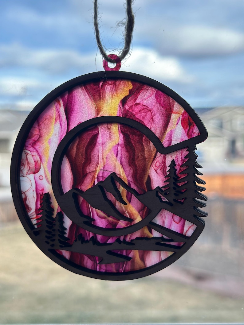 May include: A round wooden ornament with a pink and yellow abstract design. The ornament is cut out in the shape of a mountain range with trees in the foreground.