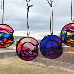 May include: Four colorful Colorado state-shaped ornaments with a mountain scene and a crescent moon. The ornaments are hanging from a window with a blurred background.