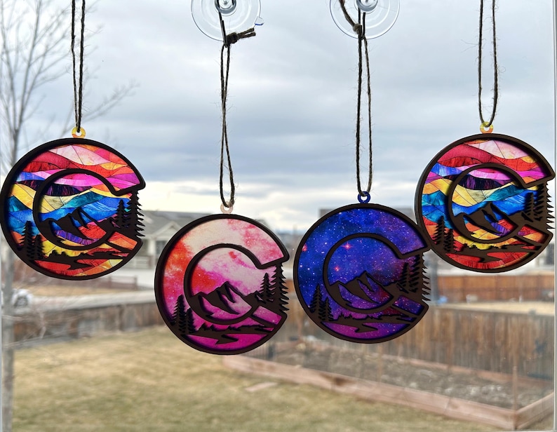 May include: Four colorful Colorado state-shaped ornaments with a mountain scene and a crescent moon. The ornaments are hanging from a window with a blurred background.