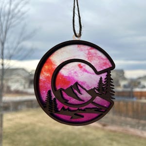 May include: A pink and orange wooden ornament shaped like the letter C with a mountain silhouette and pine trees inside. The ornament has a string for hanging.