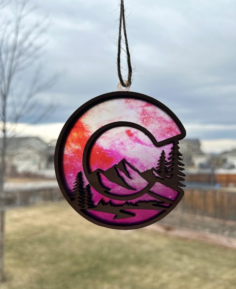May include: A pink and orange wooden ornament shaped like the letter C with a mountain silhouette and pine trees inside. The ornament has a string for hanging.