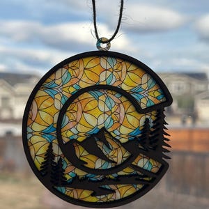 May include: A round wooden ornament with a stained glass design featuring a mountain scene and a sun. The ornament is black and has a brown string for hanging.