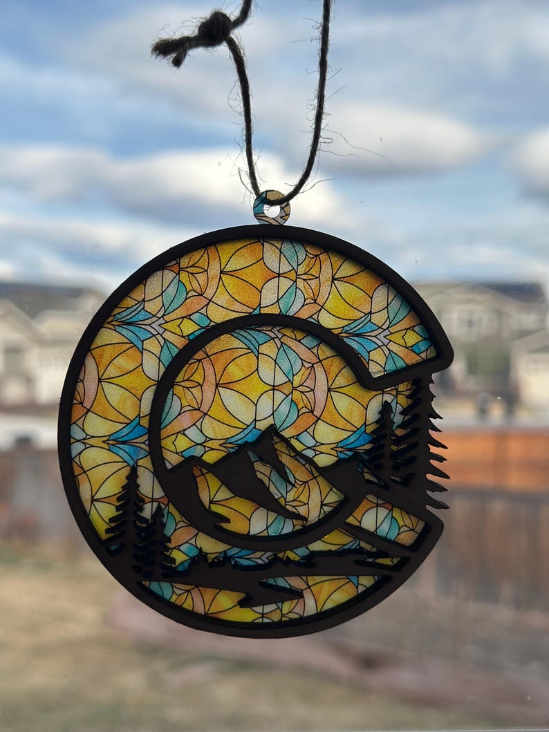 May include: A round wooden ornament with a stained glass design featuring a mountain scene and a sun. The ornament is black and has a brown string for hanging.