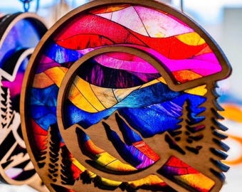 May include: A colorful wooden ornament shaped like the state of Colorado with a mountain scene inside. The ornament is painted in shades of red, orange, yellow, blue, and purple. It is hanging from a string.
