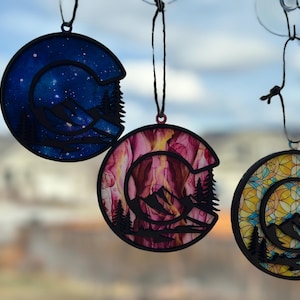 May include: Three round ornaments with a mountain silhouette design. The ornaments are made of wood and have a black outline. The first ornament has a blue and white starry sky design. The second ornament has a pink and yellow abstract design. The third ornament has a yellow and blue stained glass design.
