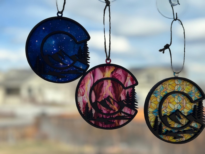 May include: Three round ornaments with a mountain silhouette design. The ornaments are made of wood and have a black outline. The first ornament has a blue and white starry sky design. The second ornament has a pink and yellow abstract design. The third ornament has a yellow and blue stained glass design.