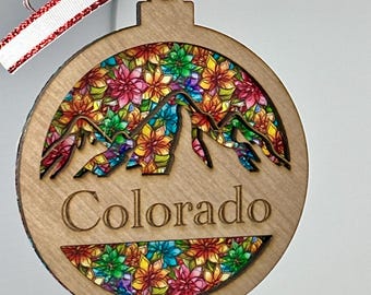 May include: Wooden Christmas ornament with a colorful floral design and the word "Colorado". The ornament features a mountain silhouette and is adorned with a red and white ribbon and a small red ball.