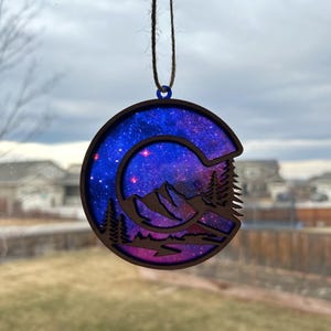 May include: A wooden ornament shaped like the letter C, with a galaxy design and a mountain scene cut out. The ornament is painted in shades of blue, purple, and pink, with stars and a crescent moon visible in the galaxy design.