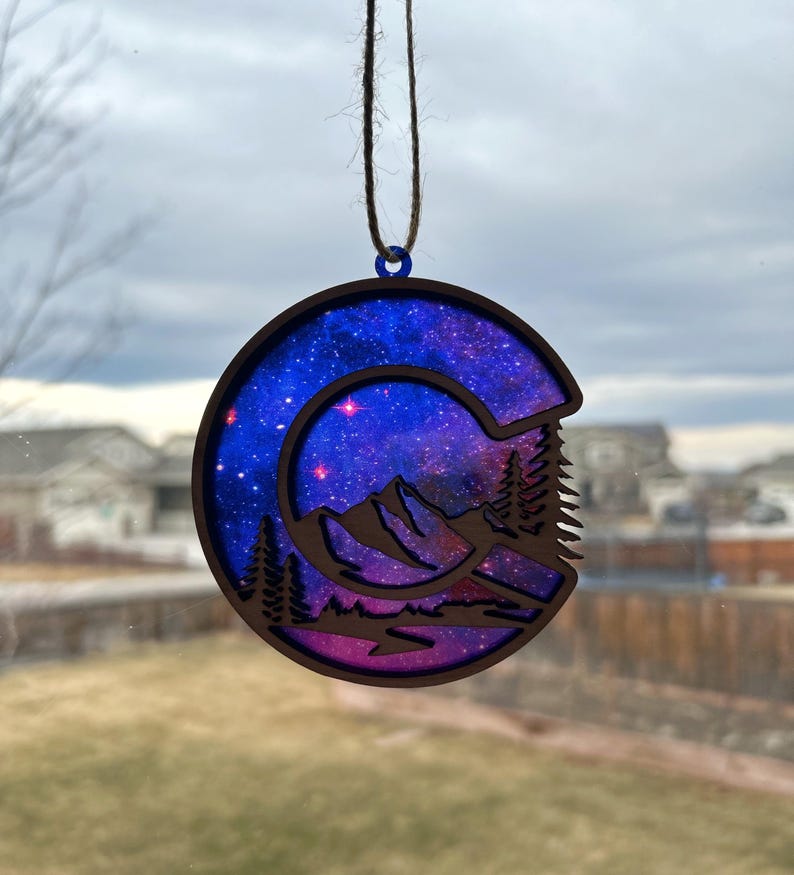 May include: A wooden ornament shaped like the letter C, with a galaxy design and a mountain scene cut out. The ornament is painted in shades of blue, purple, and pink, with stars and a crescent moon visible in the galaxy design.