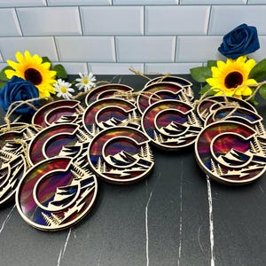 May include: A group of 12 wooden ornaments featuring a colorful mountain scene with a letter 'C' in the center. The ornaments are painted in shades of red, orange, yellow, blue, and purple. Each ornament has a string attached for hanging.