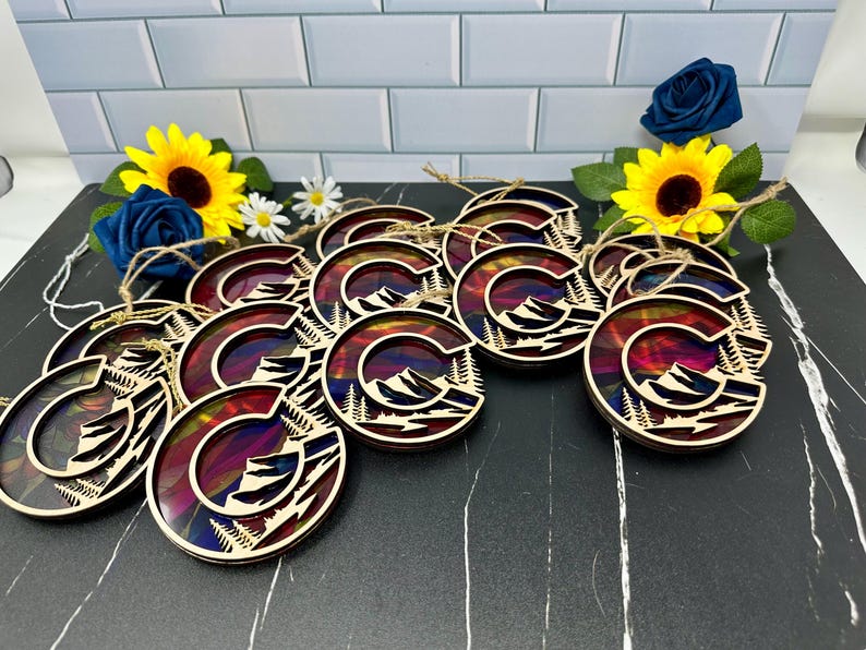 May include: A group of 12 wooden ornaments featuring a colorful mountain scene with a letter 'C' in the center. The ornaments are painted in shades of red, orange, yellow, blue, and purple. Each ornament has a string attached for hanging.
