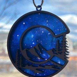 May include: A wooden ornament shaped like a mountain range with a blue and white galaxy design. The ornament is hanging from a string.