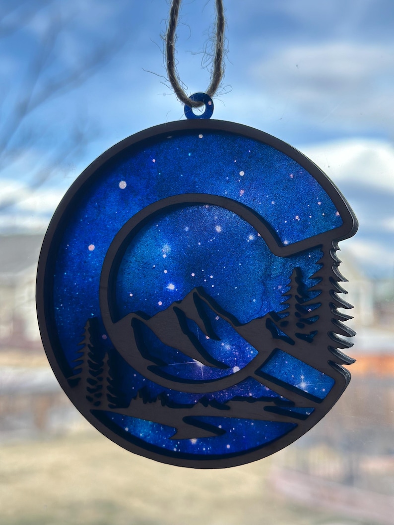 May include: A wooden ornament shaped like a mountain range with a blue and white galaxy design. The ornament is hanging from a string.