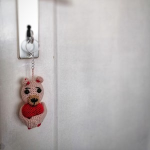May include: A pink crocheted bear holding a red heart. The bear is attached to a silver key chain and is hanging on a white door.