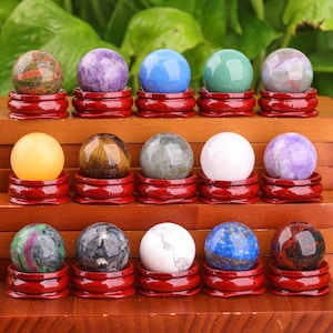 Può includere: Una collezione di 15 sfere di pietre preziose lucidate esposte su mensole di legno. Le sfere sono di vari colori, tra cui rosso, blu, verde, viola, marrone e bianco. Ogni sfera è posizionata su un piccolo supporto di legno rosso.