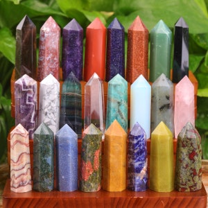 May include: A collection of twenty-one polished gemstone obelisks in various colors, including amethyst, rose quartz, and green aventurine. The obelisks are displayed on a wooden stand against a backdrop of green foliage.