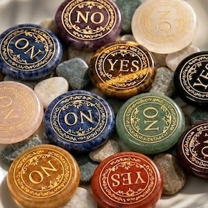 May include: A collection of circular polished stones in various colours including purple, blue, green, orange, red, black, and translucent white, each engraved with the word YES or NO in gold lettering, arranged on a square white ceramic tray.