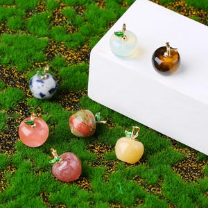 May include: A selection of small, apple-shaped gemstone pendants in shades of pink, yellow, blue, and brown. Each pendant features a gold-coloured stem and leaf detail. The pendants are presented on a bed of green moss.
