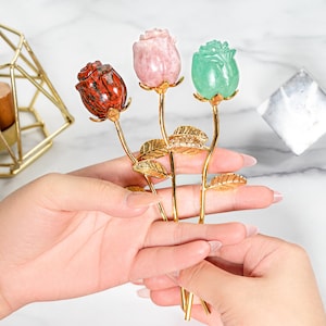 May include: Three gemstone rose sculptures in red, pink, and green. Each rose has a gold-coloured stem and leaves. The roses are held in a person's hands, with a marble background.