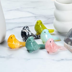 May include: A collection of small, colorful bird figurines in various colors, including yellow, green, blue, pink, and black and white. The birds are made of a glossy material and are arranged on a white marble surface.