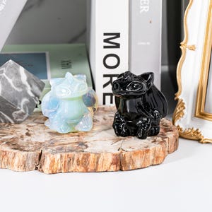 May include: Two dragon figurines, one opalescent and the other black, sit on a wooden slab. The opalescent dragon has a light blue and white color scheme, while the black dragon is glossy. The figurines are detailed and have wings.