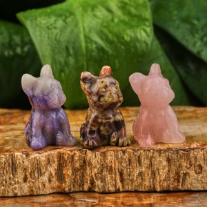May include: Three small carved stone figurines of dogs in different colors. The dog on the left is purple, the middle dog is brown and black, and the dog on the right is pink.