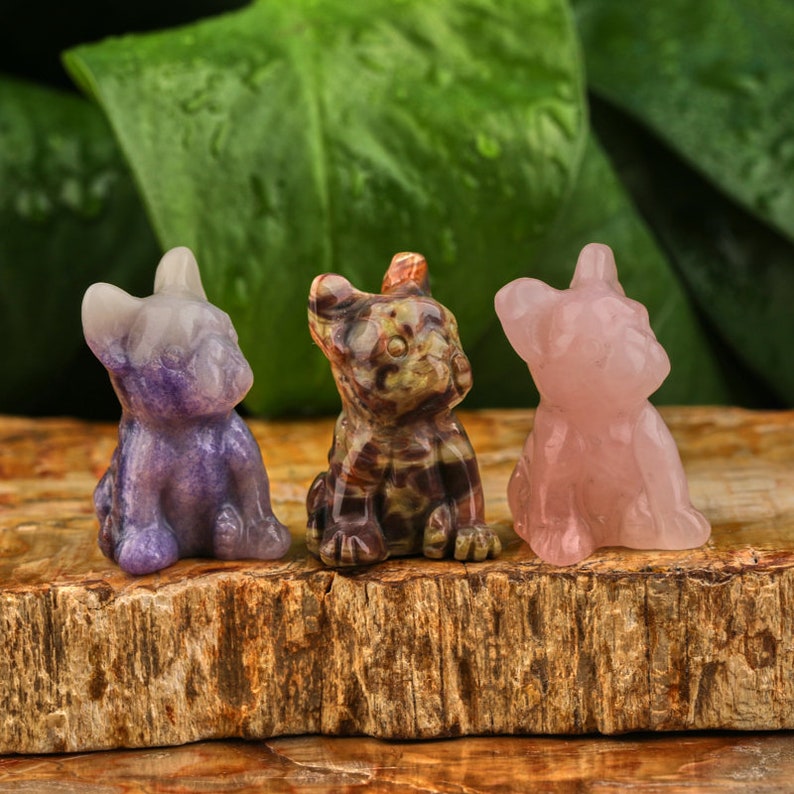 May include: Three small carved stone figurines of dogs in different colors. The dog on the left is purple, the middle dog is brown and black, and the dog on the right is pink.