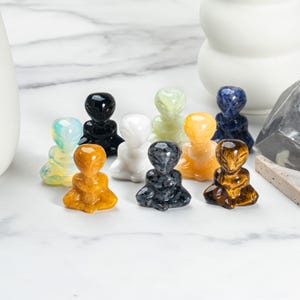 May include: A selection of small alien figurines in a variety of colours, including yellow, black, white, blue, and green. The figurines are made of polished stone and are arranged on a white marble surface.
