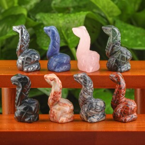 May include: Eight small carved stone snake figurines in various colors, including black, gray, blue, pink, red, and green. The figurines are arranged in two rows on a wooden surface.