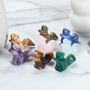 May include: A collection of small, carved squirrel figurines in various colors and patterns. The squirrels are made from different types of polished gemstones, including amethyst, rose quartz, and tiger's eye. The figurines are displayed on a white surface.