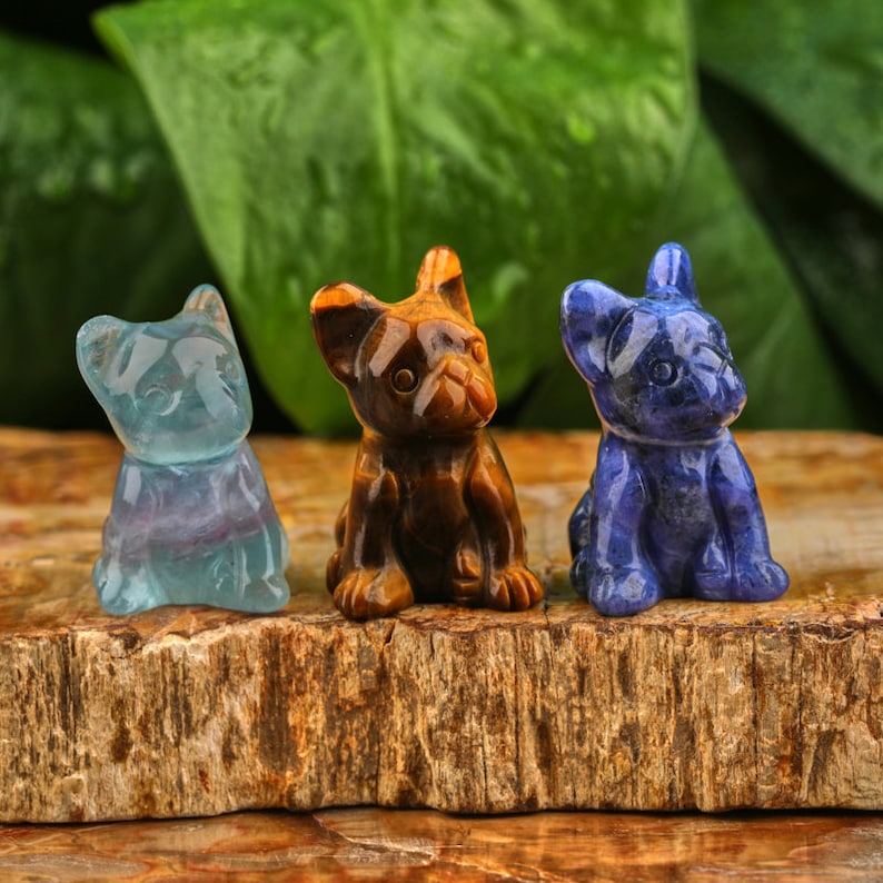 May include: Three small carved dog figurines made of tiger's eye, blue lace agate, and fluorite. The figurines are sitting on a wooden surface.