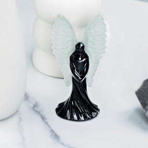 May include: A black angel figurine with translucent white wings. The angel is wearing a long, flowing robe. The figurine is on a white marble surface. The angel is a decorative item.