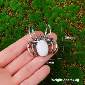 May include: A silver spider ring featuring a white oval stone. The ring is 33mm wide and 36mm long, weighing approximately 8g. The ring is displayed against a green backdrop.