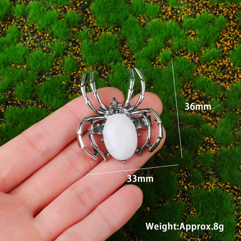 May include: A silver spider ring featuring a white oval stone. The ring is 33mm wide and 36mm long, weighing approximately 8g. The ring is displayed against a green backdrop.