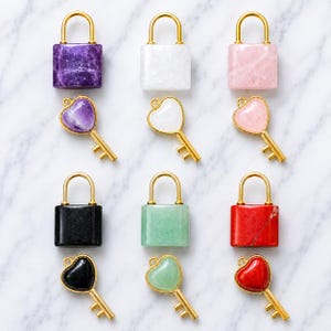 May include: A collection of six rectangular stone padlocks and six matching heart-shaped keys, each featuring a gold-tone metal frame. The stones include purple, white, pink, black, green, and red varieties, arranged in two rows of three pairs.