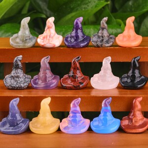 May include: A collection of 15 witch hat shaped crystal carvings in various colours, including black, white, purple, pink, red, orange, blue, and green. The hats are arranged in three rows on a wooden surface.