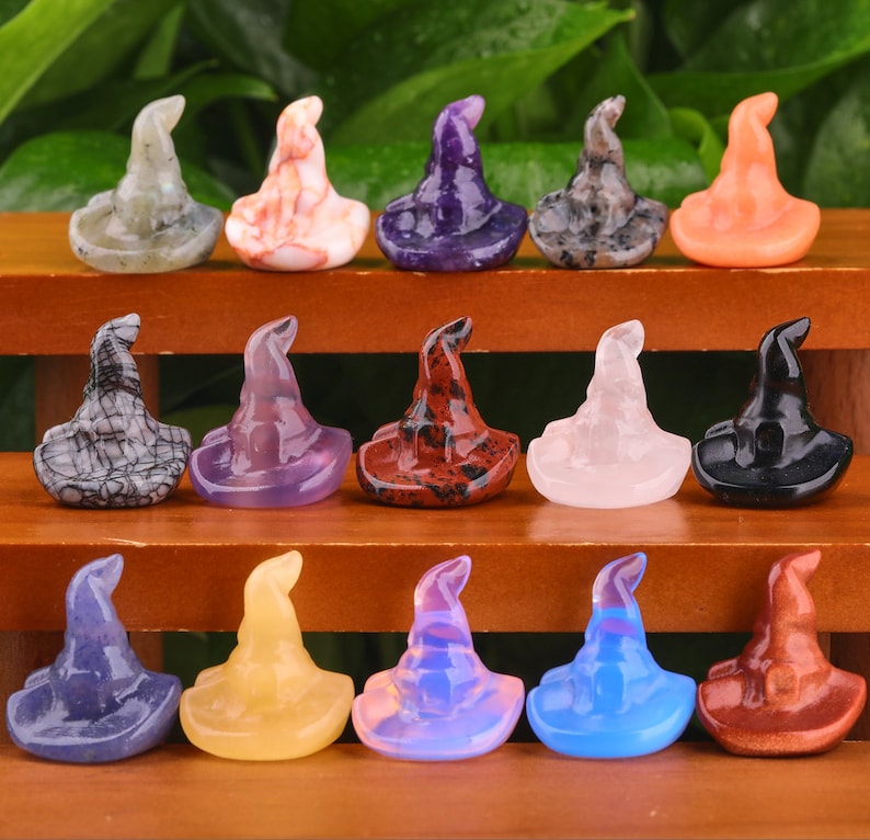 May include: A collection of 15 witch hat shaped crystal carvings in various colours, including black, white, purple, pink, red, orange, blue, and green. The hats are arranged in three rows on a wooden surface.