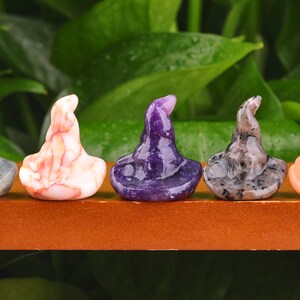 May include: Five miniature witch hats crafted from various types of stone. The hats are displayed in a row on a wooden shelf. The hats are made from labradorite, pink and white agate, amethyst, granite, and orange agate.