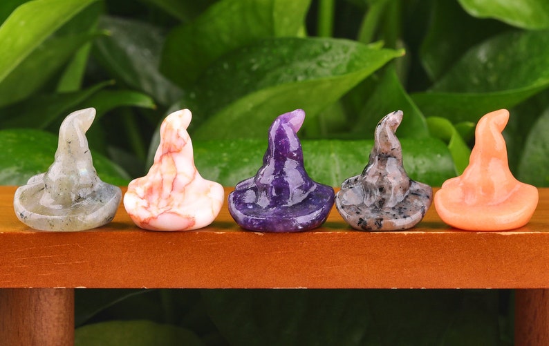 May include: Five miniature witch hats crafted from various types of stone. The hats are displayed in a row on a wooden shelf. The hats are made from labradorite, pink and white agate, amethyst, granite, and orange agate.
