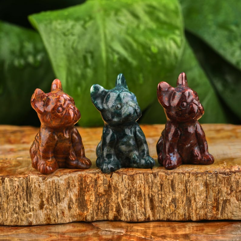May include: Three small carved stone figurines of French Bulldogs. The figurines are in a row on a wooden surface. The dog on the left is brown, the middle dog is green and black, and the dog on the right is red.