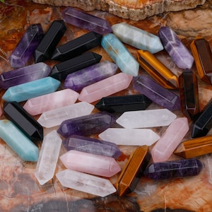 Könnte beinhalten: Eine Sammlung von Kristallstäben in verschiedenen Farben, darunter Amethyst, Rosenquarz, klarer Quarz, Tigerauge und schwarzer Obsidian.