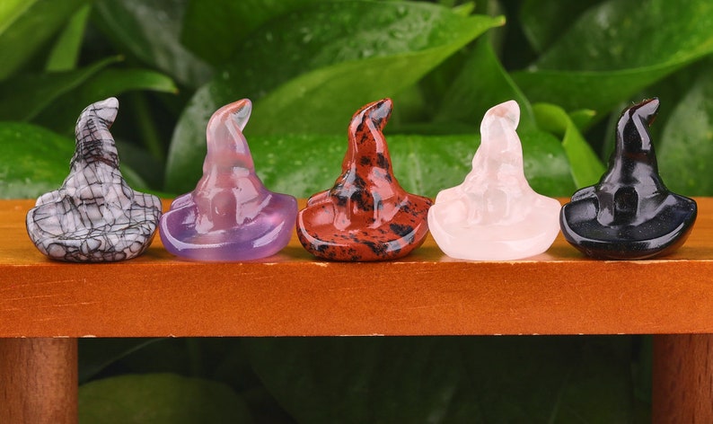 May include: Five miniature witch hats crafted from various types of stone. The hats are displayed in a line on a wooden surface. The hats are black, white, purple, red, and pink.