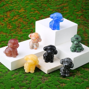 May include: A selection of small gemstone dog figurines in colours such as red, blue, black, yellow, and green. The figurines are displayed on white platforms and a textured surface against a green background.
