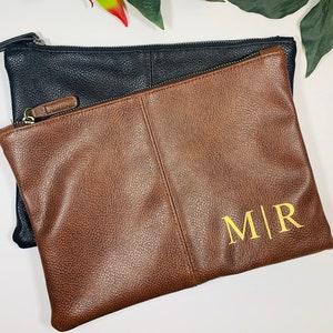 Könnte beinhalten: Eine braune Leder-Clutch mit einem schwarzen Leder-Reißverschluss. Die Clutch hat ein goldfarbenes, geprägtes Monogramm "M|R" auf der Vorderseite.