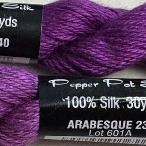 May include: Two skeins of purple Pepper Pot Silk embroidery thread. Each skein is labeled 100% Silk 30yds. The top label reads PIROUETTE 240 Lot 512A, and the bottom label reads ARABESQUE 239 Lot 601A. The thread has a visible sheen and texture.