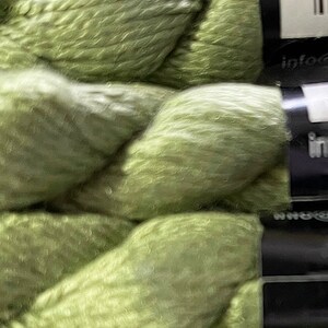 May include: Close-up of several skeins of light green yarn. The yarn is tightly wound and has a soft, textured appearance. The image highlights the color and texture of the yarn, suitable for knitting or crochet projects.